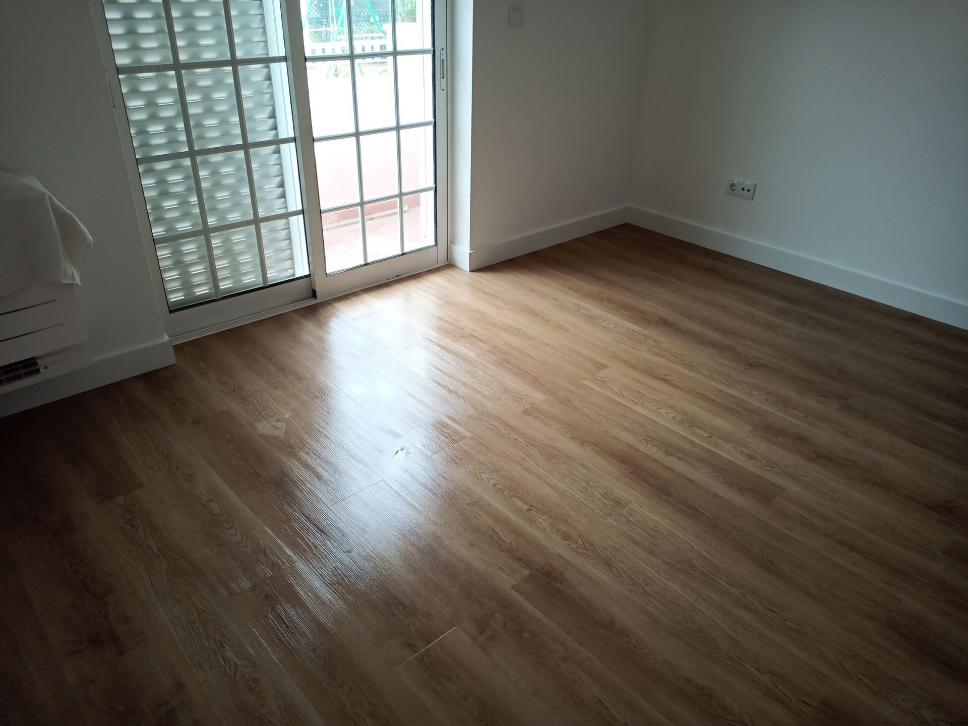 Quarto vazio com piso de madeira, paredes brancas e porta de vidro.