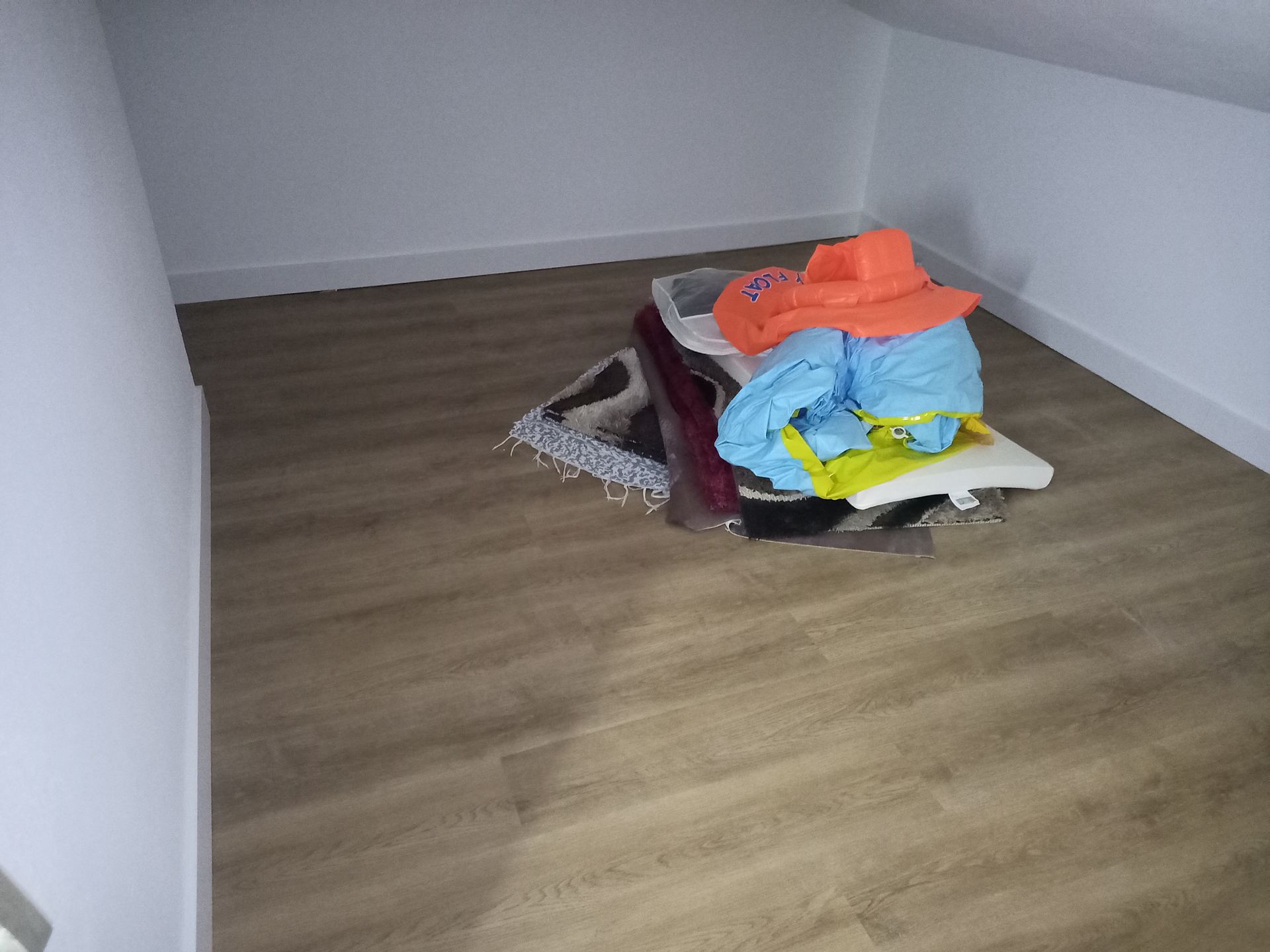Quarto pequeno com piso imitando madeira, paredes brancas e uma pilha de roupas coloridas.
