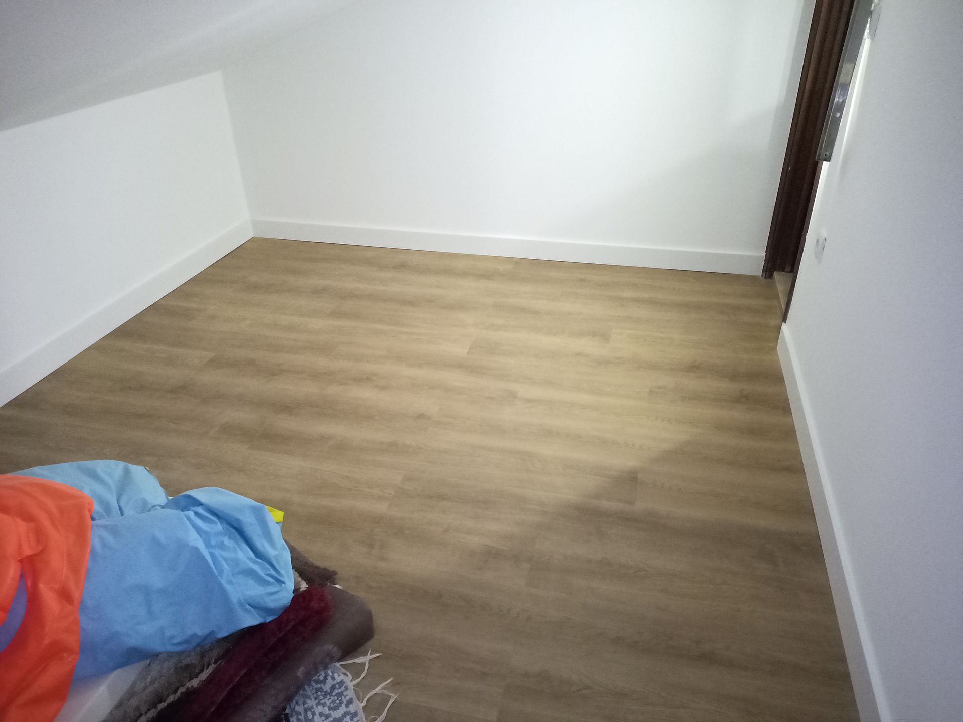 Quarto vazio com piso que imita madeira clara, paredes brancas e uma porta à direita. Uma pilha de roupas coloridas está em primeiro plano.