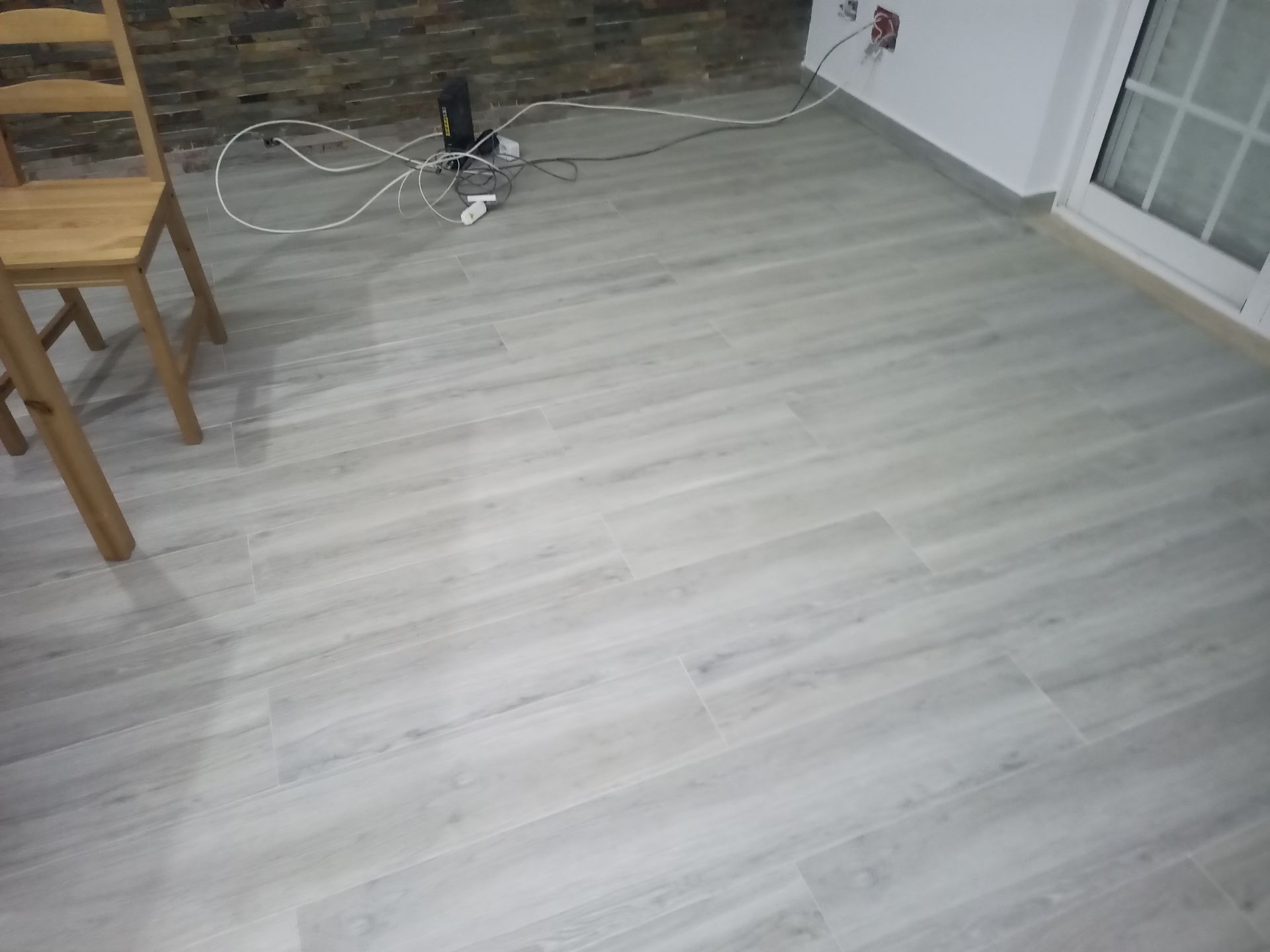 Piso imitando madeira em tom cinza claro em um cômodo com uma cadeira de madeira e alguns cabos.