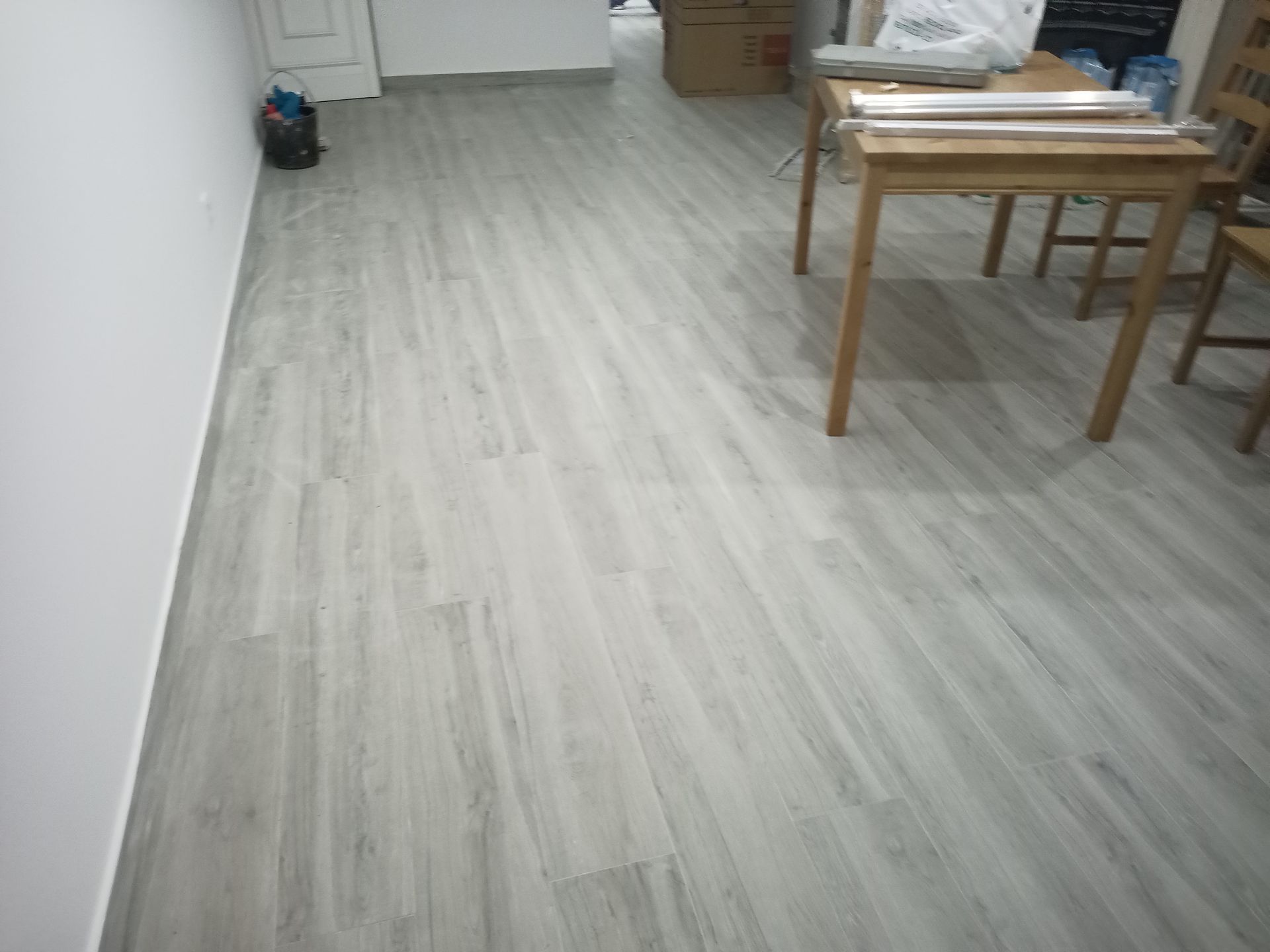 O piso que imita madeira em tom cinza claro se estende por toda a sala. Uma mesa e cadeiras de madeira estão ao fundo.