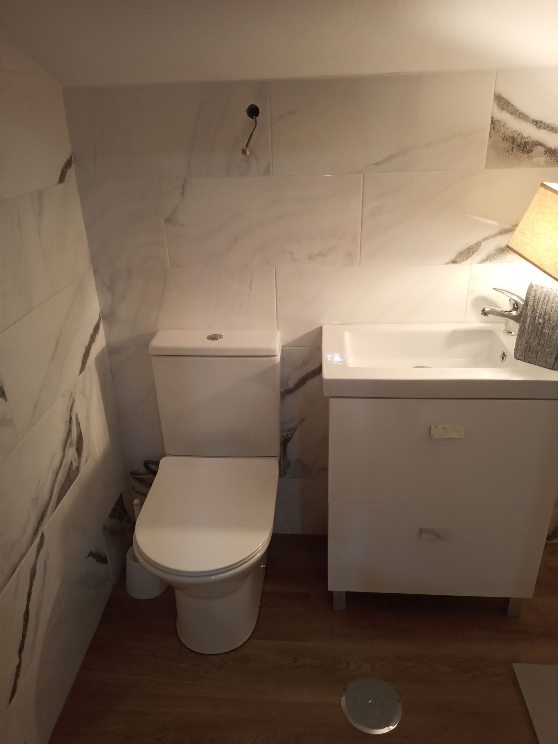 Banheiro pequeno com vaso sanitário branco, pia e paredes revestidas com azulejos que imitam mármore. Piso de madeira.