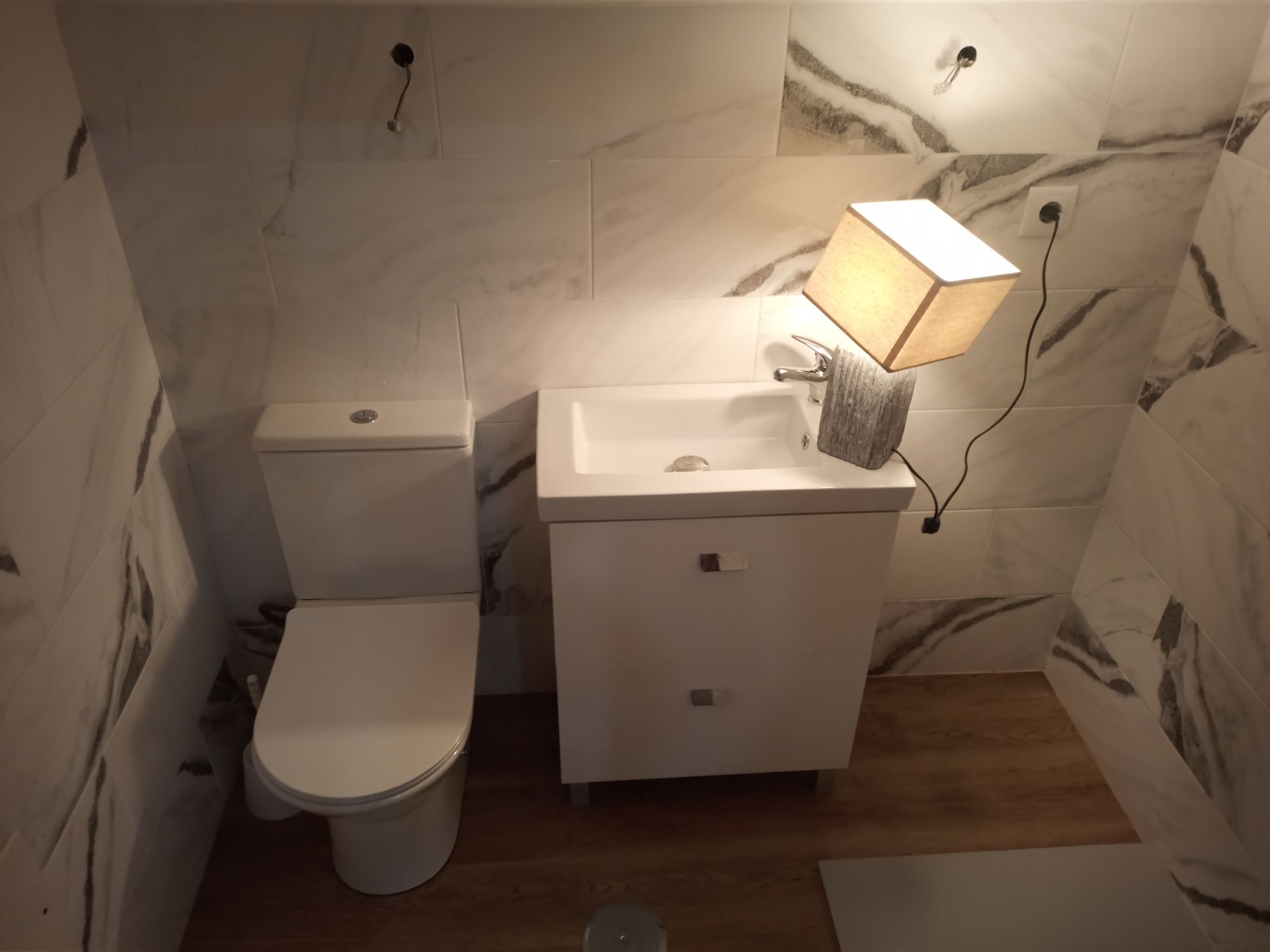 Banheiro com vaso sanitário, pia e luminária brancos. Paredes com estampa de mármore.