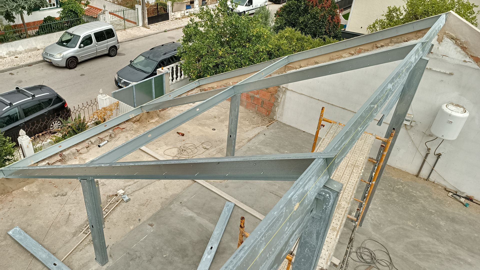 Estrutura metálica de um telhado em construção em uma casa, com veículos estacionados nas proximidades.