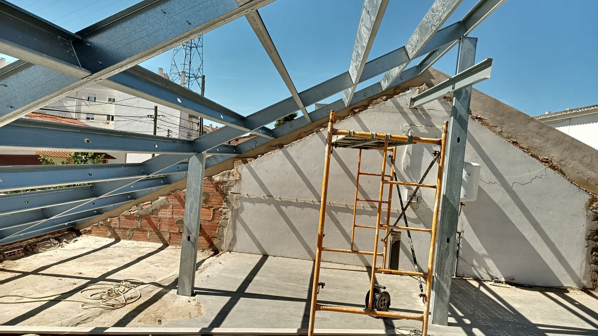Vigas metálicas compõem a estrutura do telhado sobre um espaço exterior; um andaime amarelo está posicionado no centro.