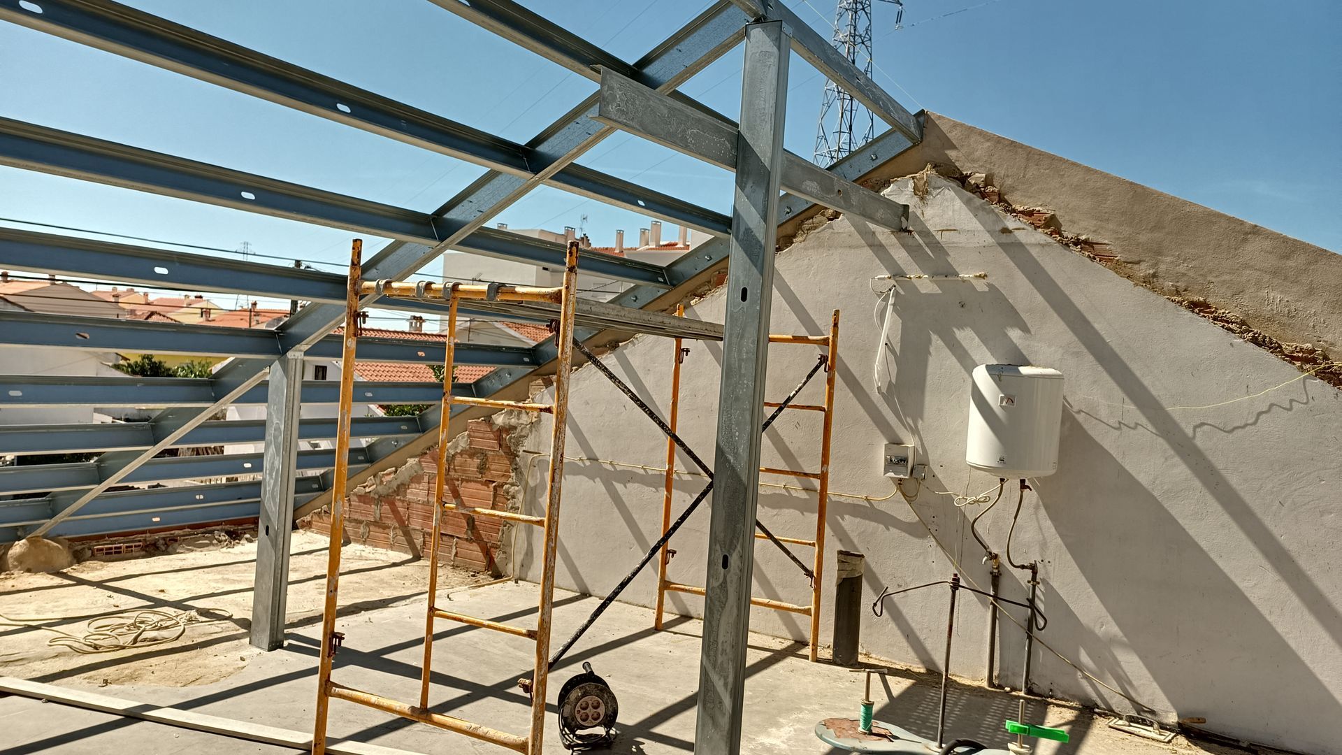 Estrutura metálica de uma construção em andamento ao lado de uma parede branca sob um céu azul. Andaimes já estão instalados.