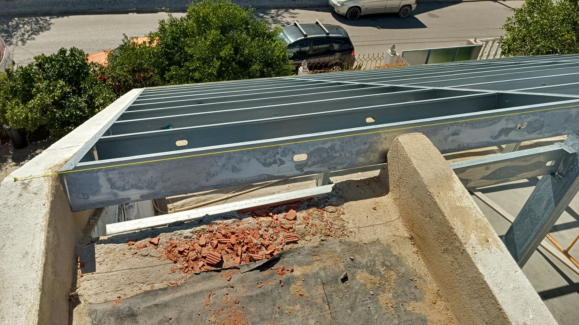 Estrutura metálica do telhado sobre uma base de concreto com entulho; árvores e carros visíveis ao fundo.