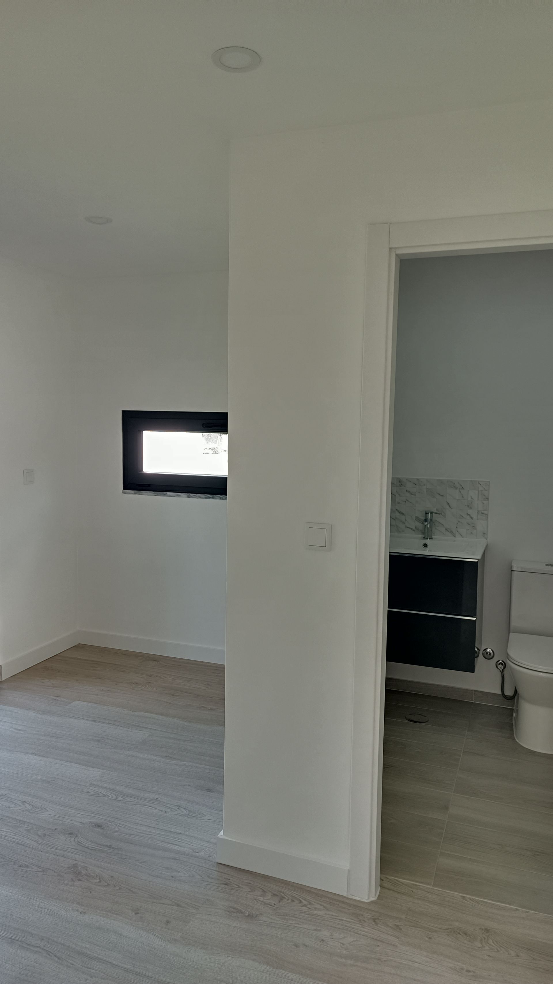 Vista interior de um quarto moderno com janela, porta e banheiro visíveis. As paredes são brancas e o piso é cinza claro.