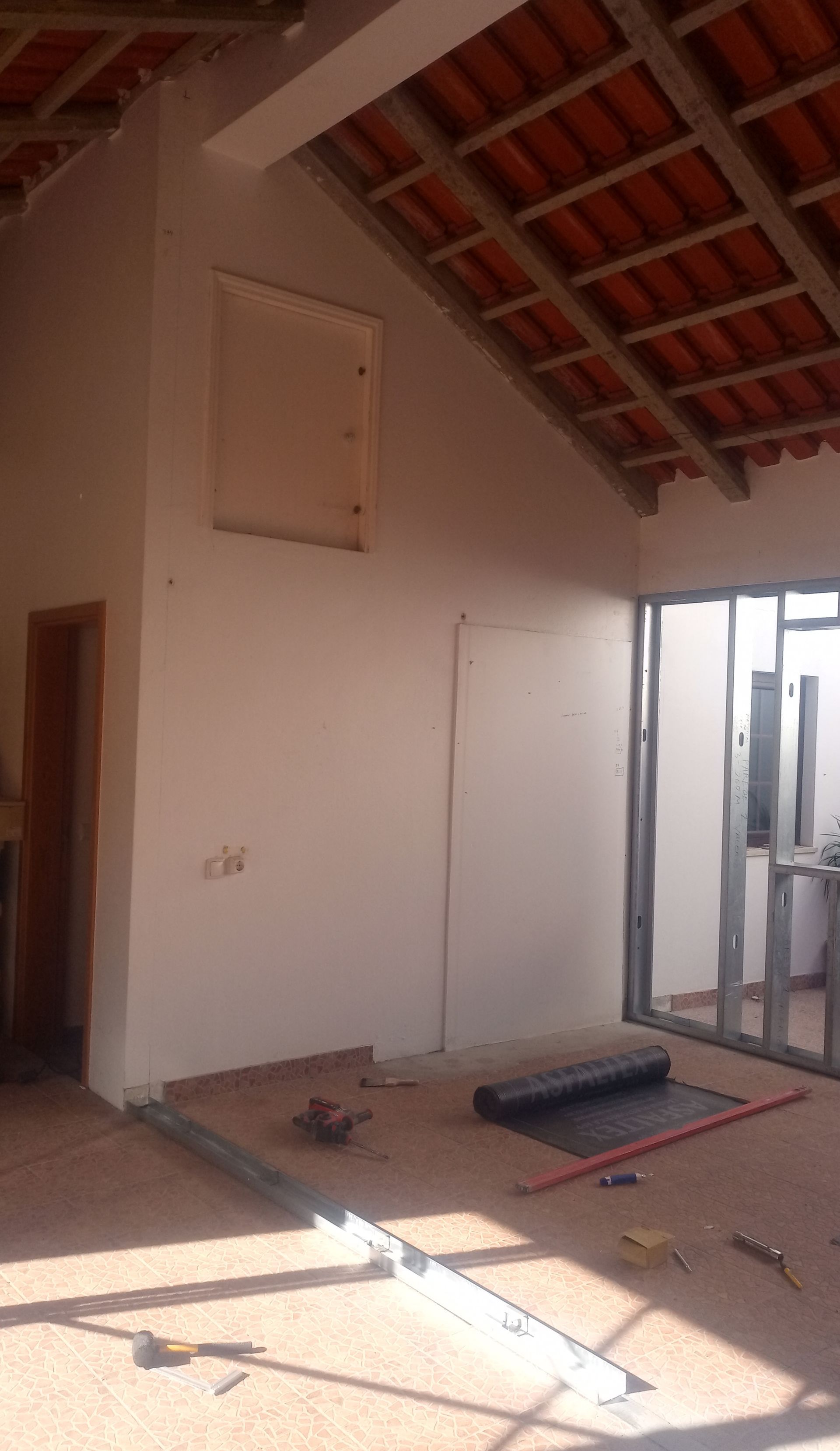Interior de um cômodo em construção com paredes brancas, telhas aparentes e estrutura metálica.