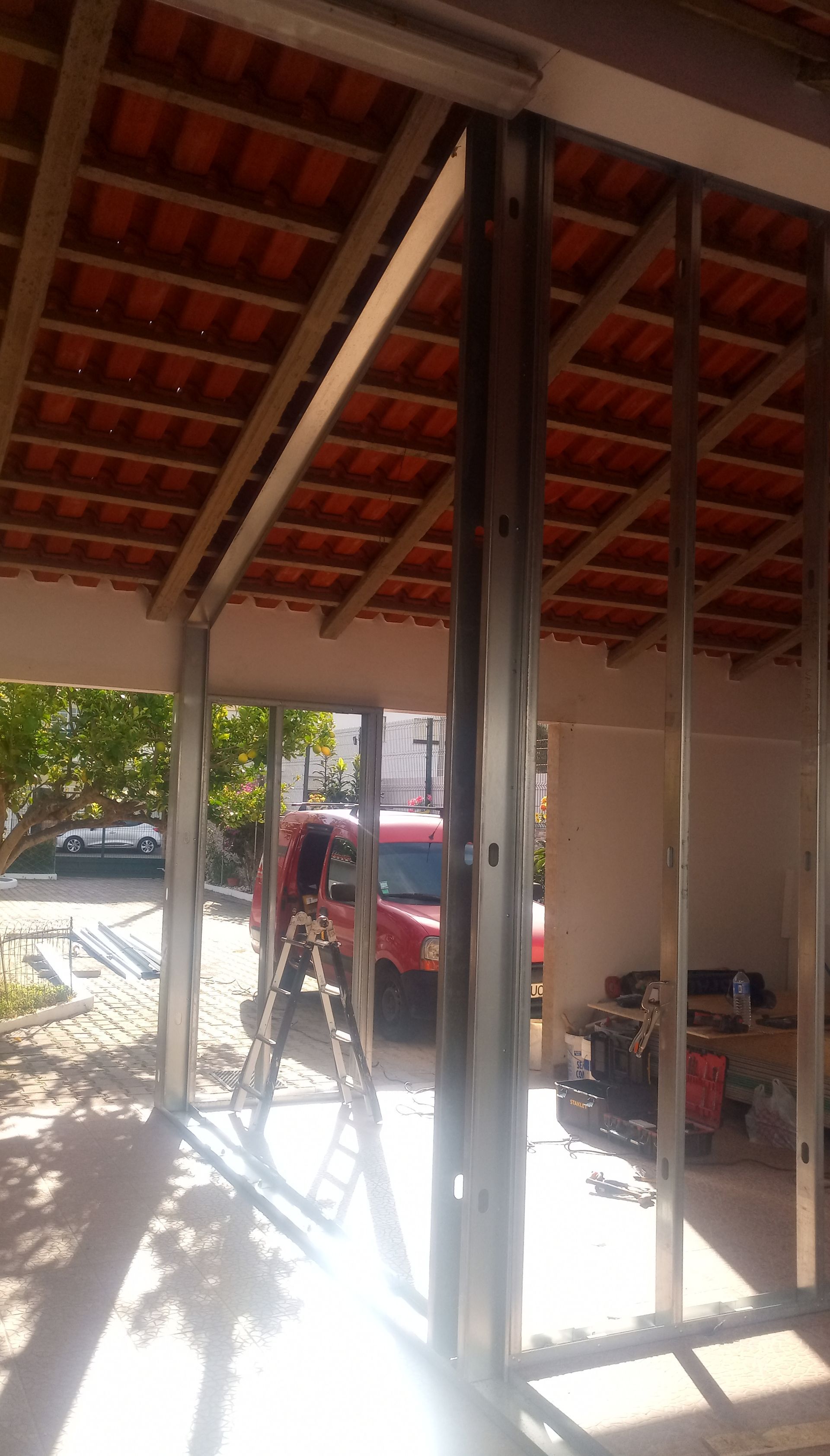 Estrutura metálica de uma construção em andamento; telhado de telhas vermelhas visível; escada encostada dentro da estrutura.