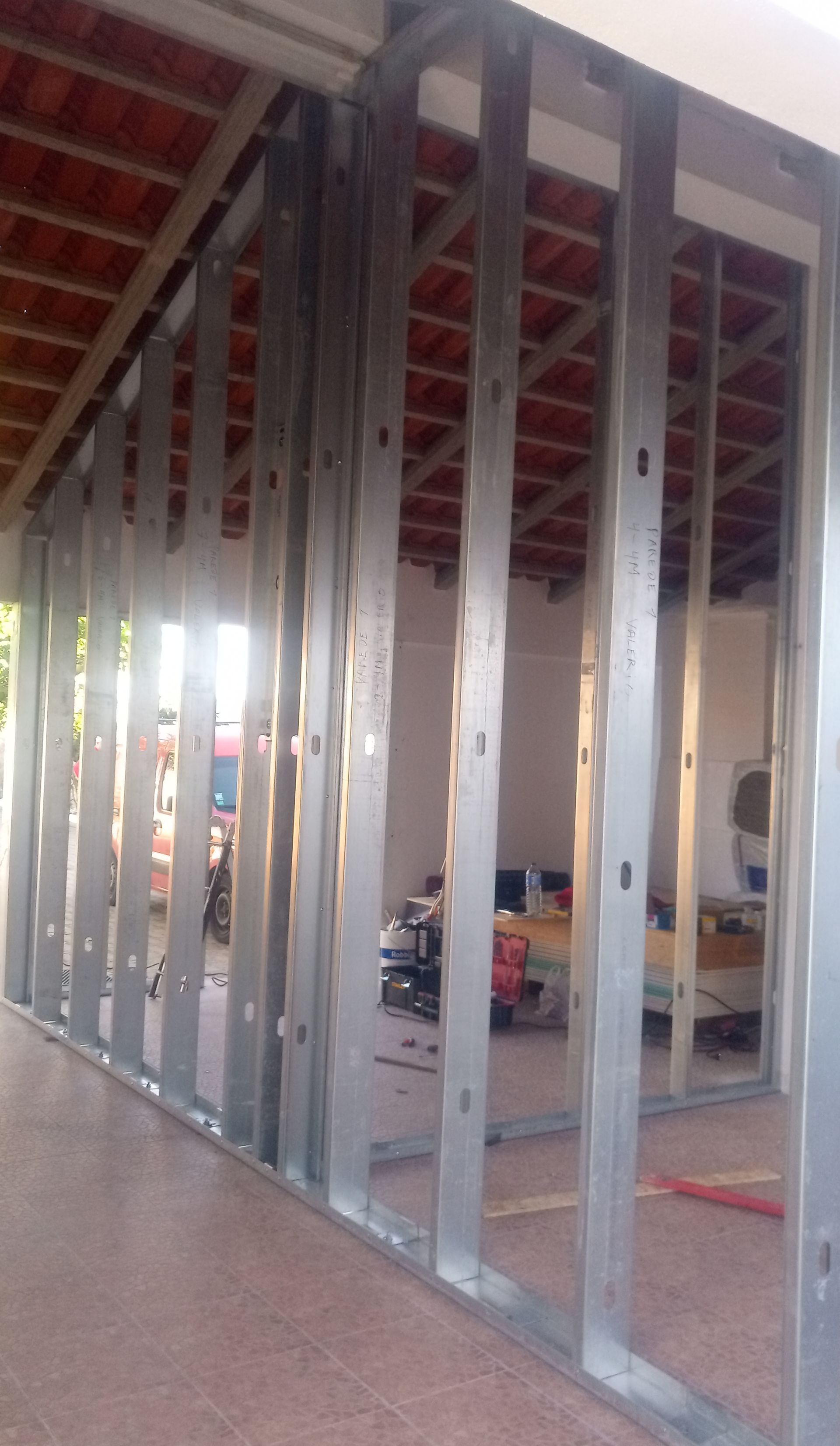 Estrutura metálica de uma parede em construção, com um telhado de telhas vermelhas ao fundo.