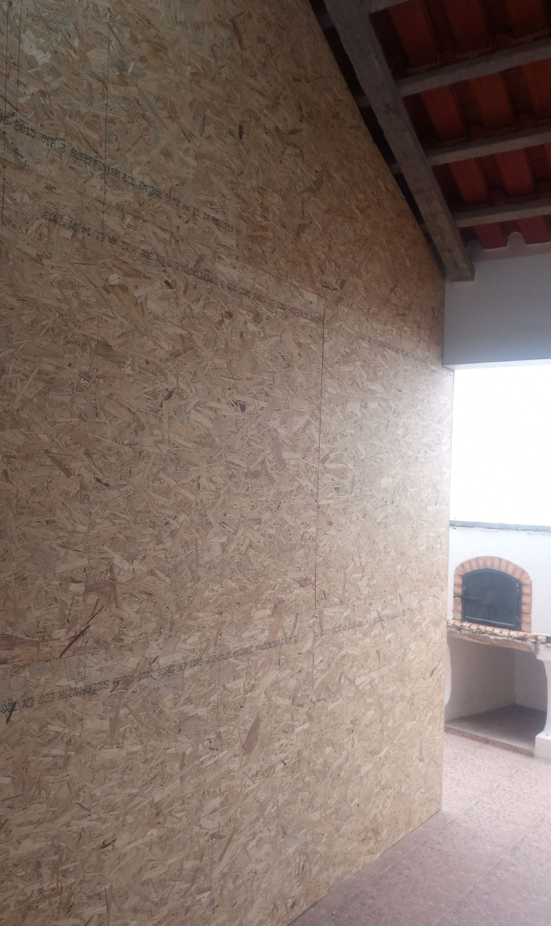 Parede feita de painéis de madeira, adjacente a uma estrutura externa de tijolos, sob um telhado de telhas de terracota.