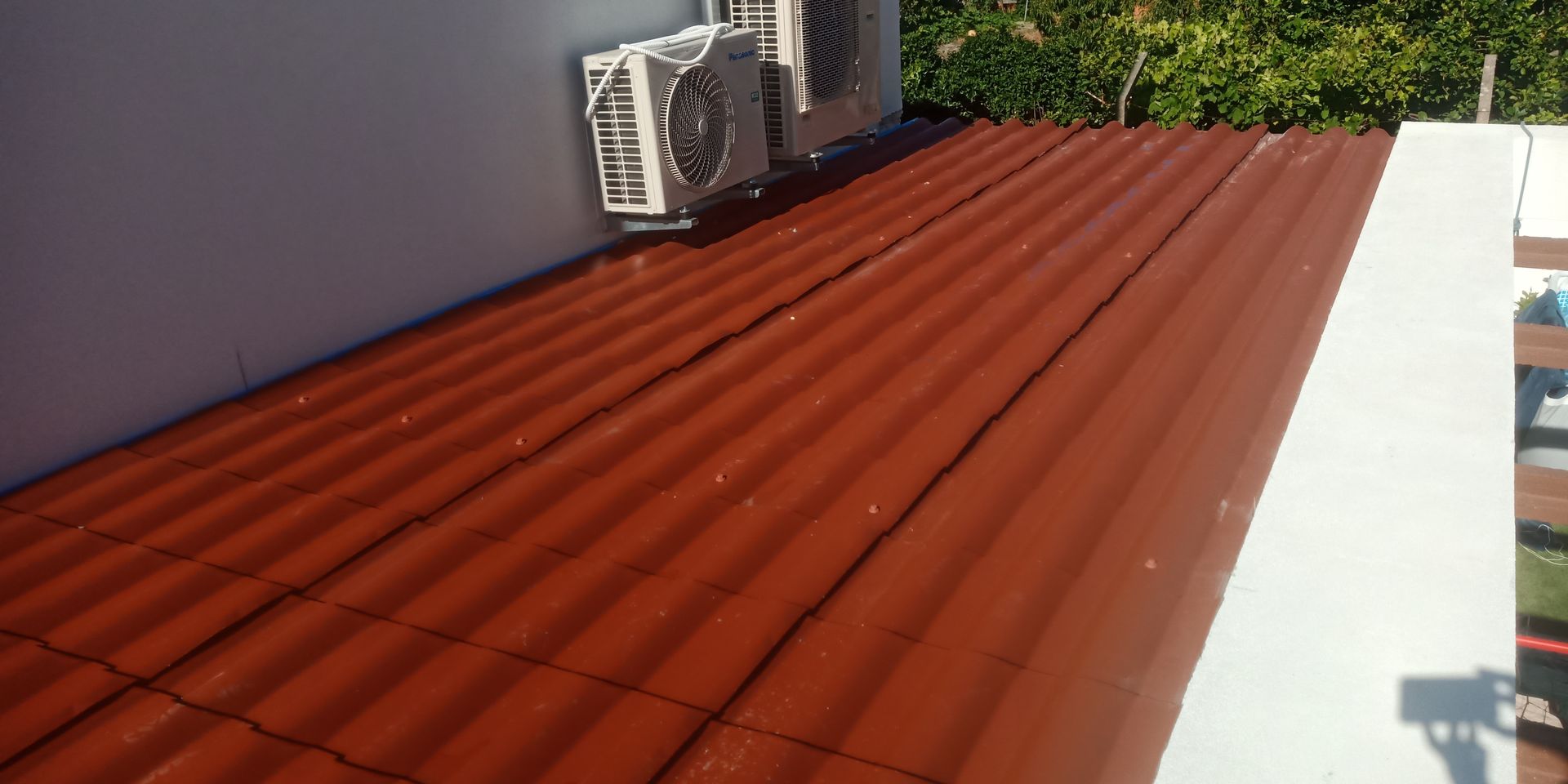 Telhado de metal ondulado vermelho ao lado de uma parede branca, com aparelhos de ar condicionado instalados na parede.