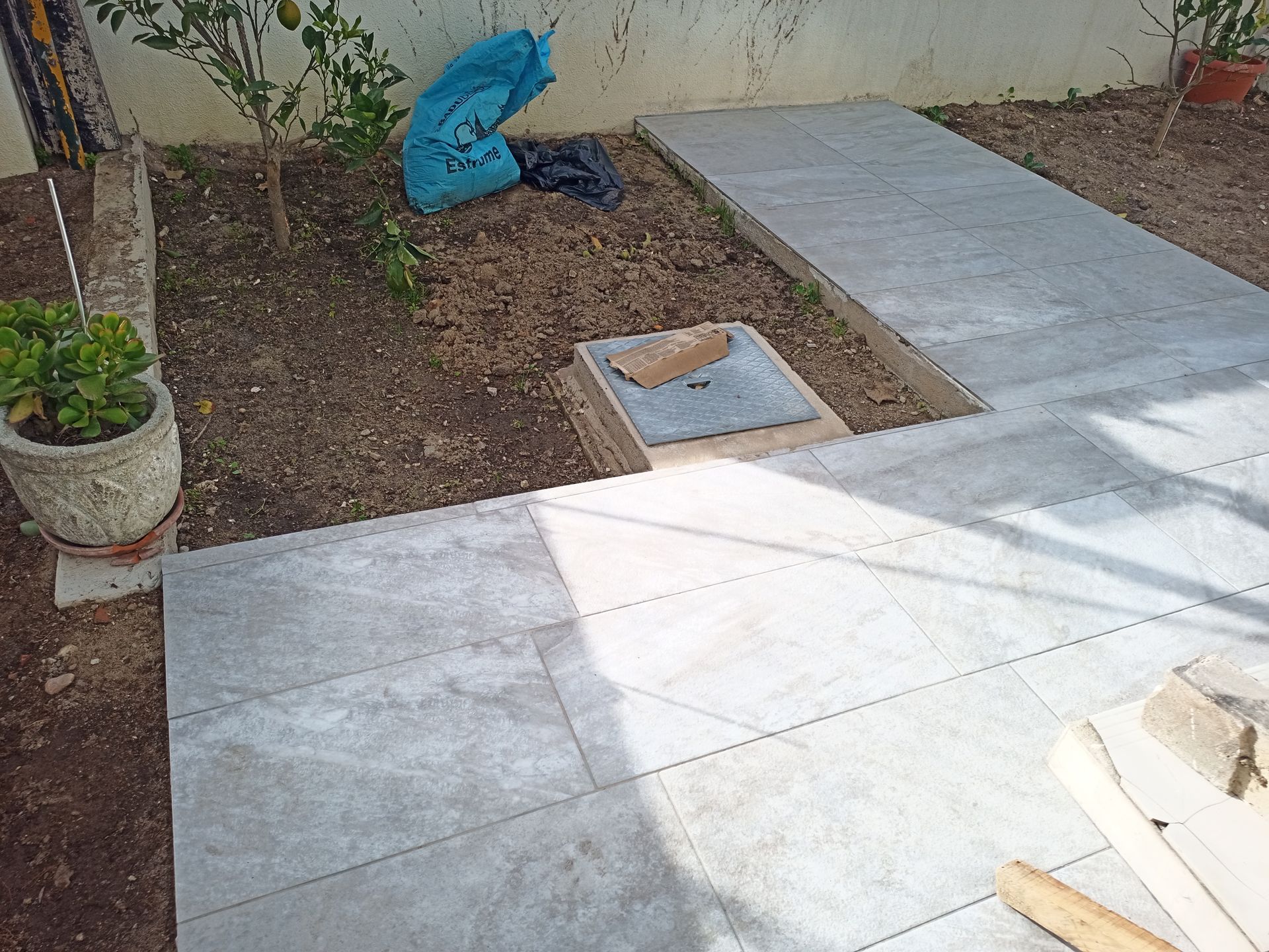 Pedras de pavimentação cinzentas sendo instaladas em um jardim, com solo exposto e um vaso de planta próximo.
