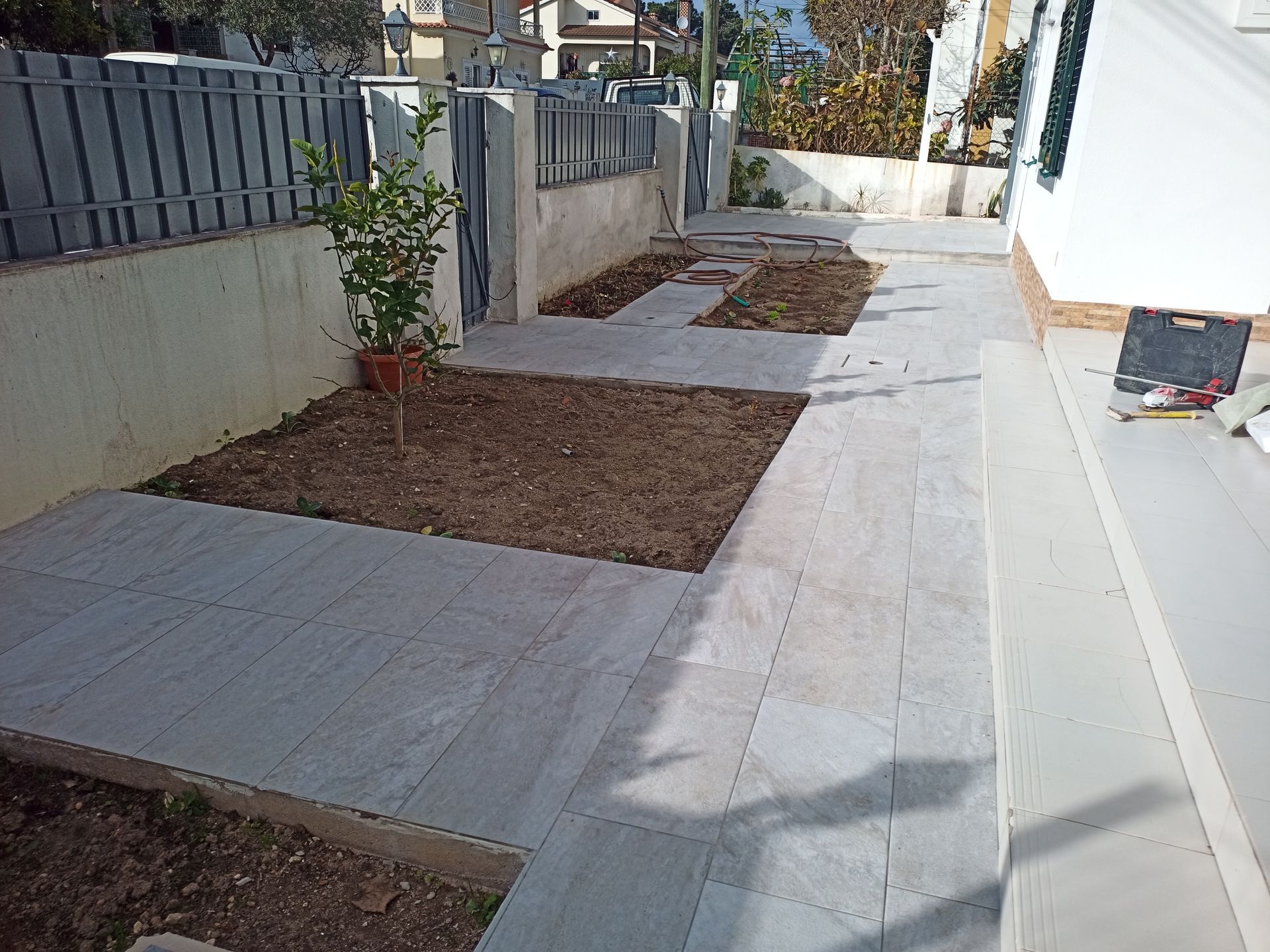Caminho pavimentado com canteiros de terra, uma pequena árvore e uma cerca ao lado de um edifício.