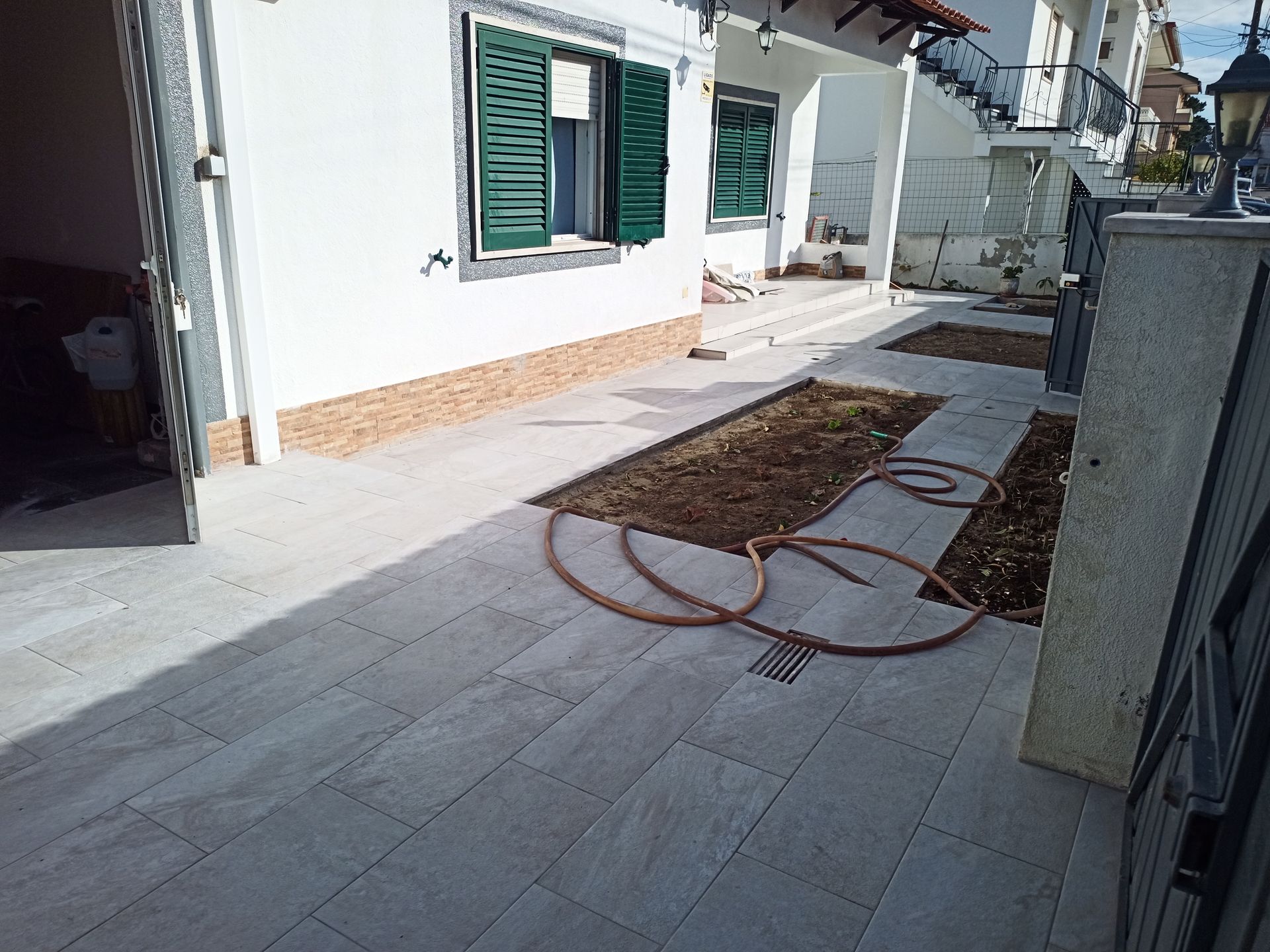 Exterior de um prédio branco com venezianas verdes, caminho de ladrilhos cinza e canteiros de jardim com mangueira.