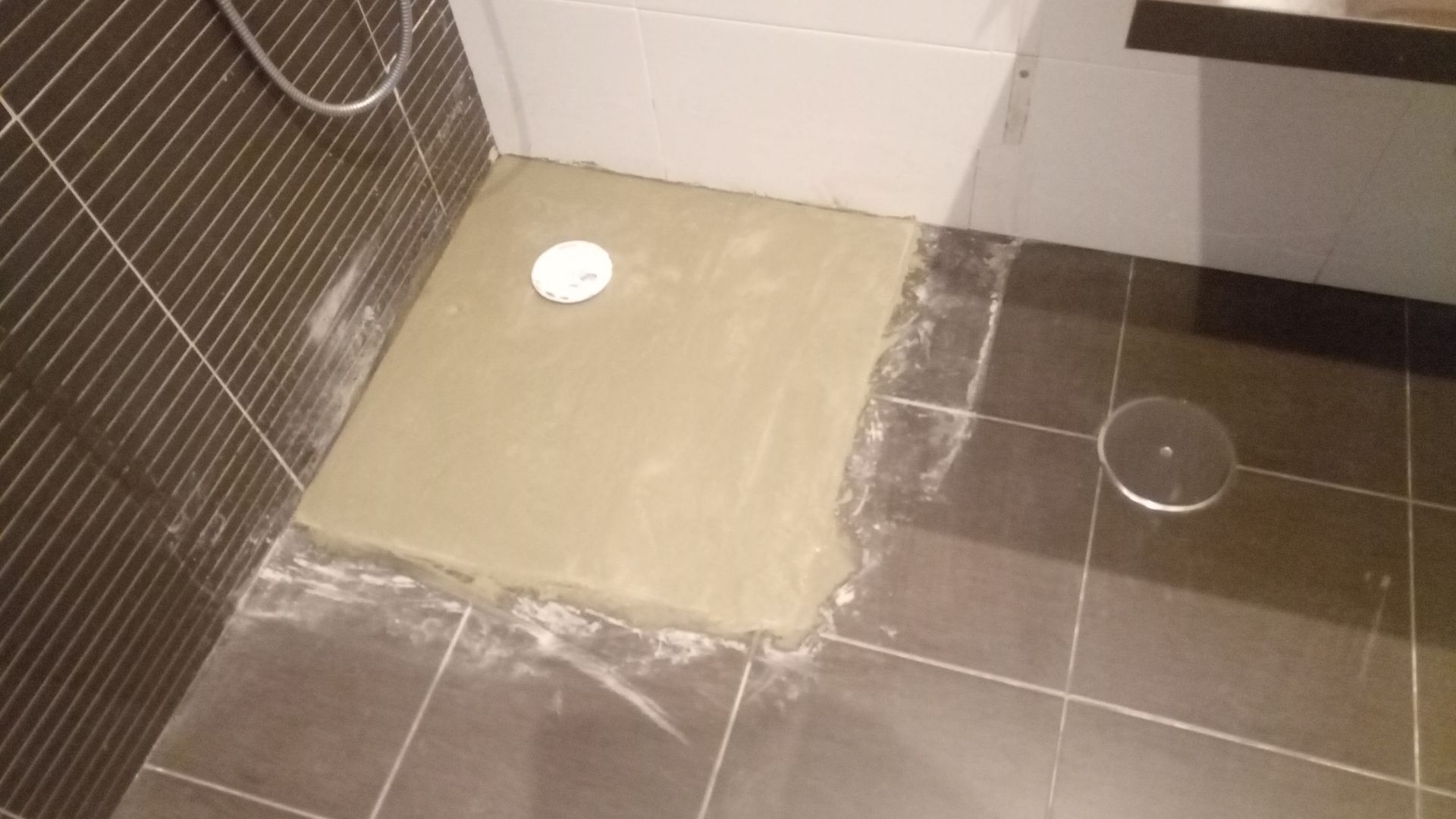 Piso do banheiro parcialmente coberto com cimento fresco, próximo ao ralo. Azulejos escuros contornam o material novo.
