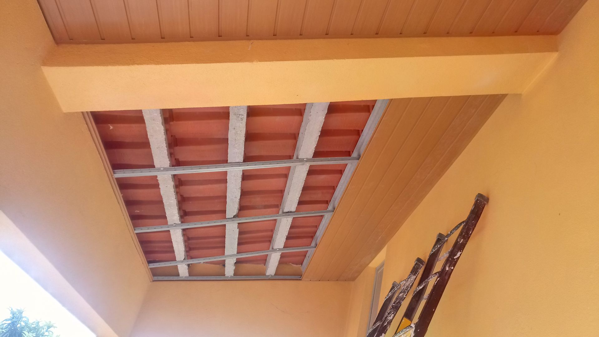 Teto pintado de laranja com telhas vermelhas expostas e vigas de madeira. Uma escada encostada em uma parede amarela.