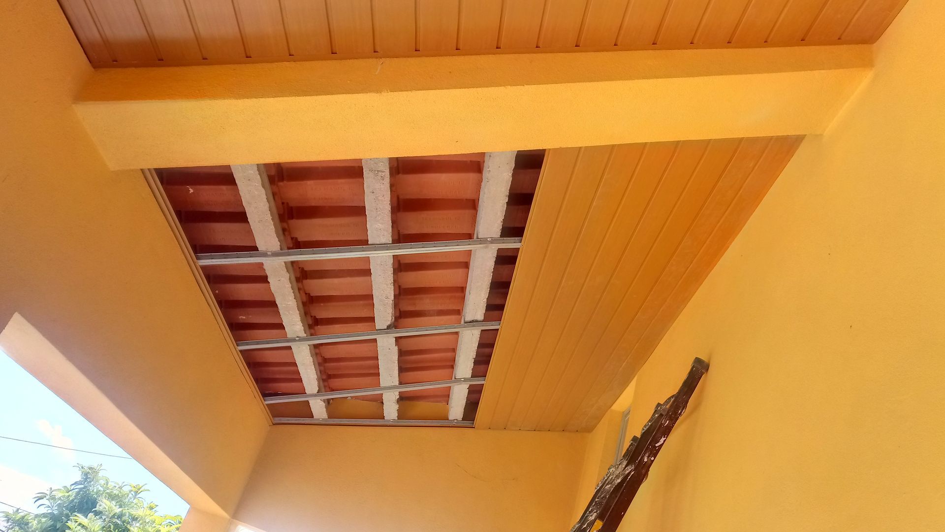 Teto amarelo da varanda com tábuas de madeira, telhas vermelhas expostas e vigas estruturais.