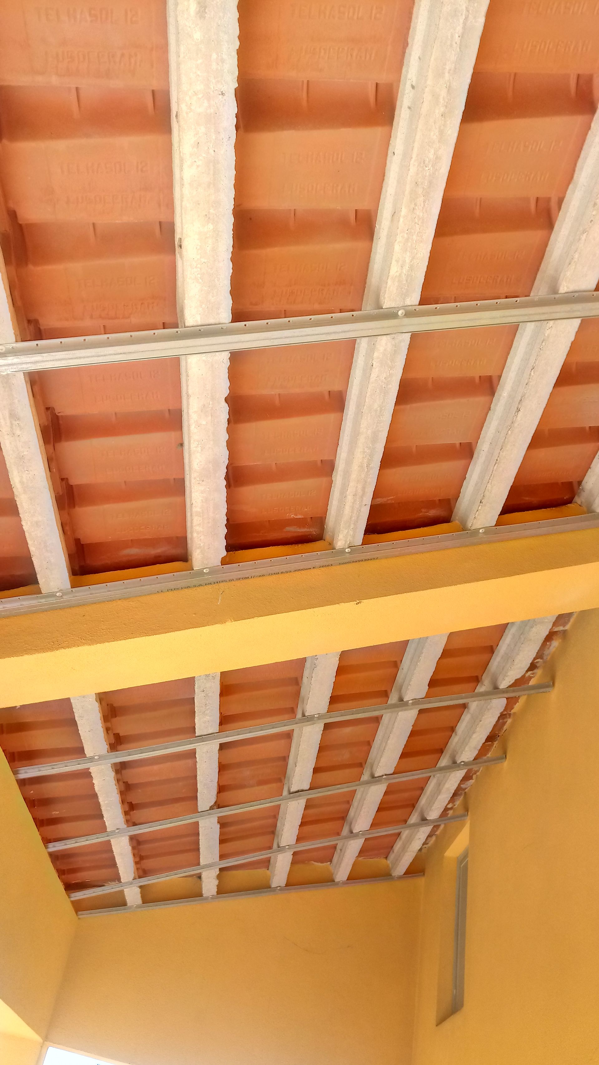 Vista de baixo para cima, mostrando um telhado de telhas de terracota com vigas de concreto e uma parede amarela em um canteiro de obras.