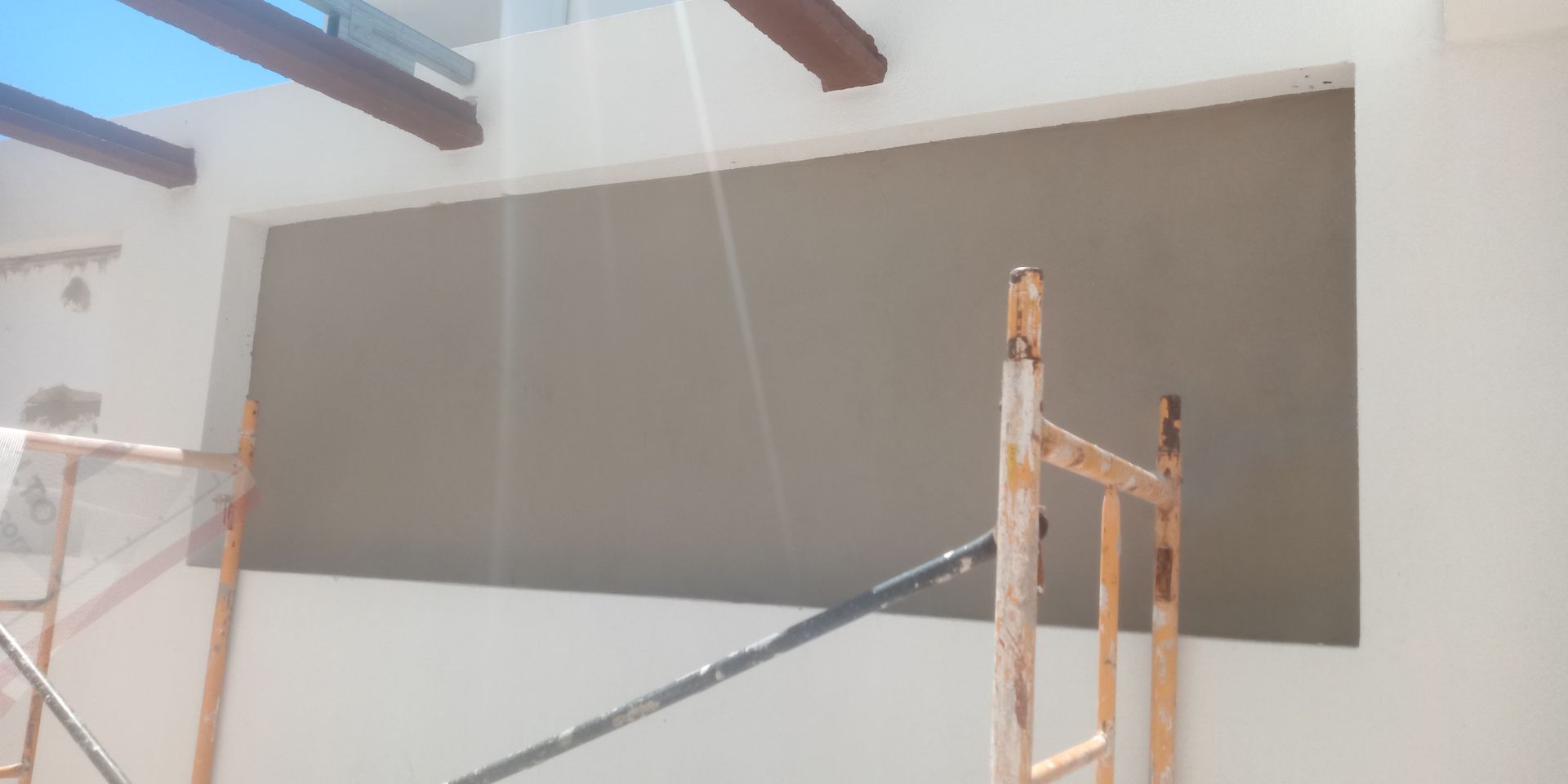 Canteiro de obras com andaimes e uma grande janela vazia; vigas marrons no teto.