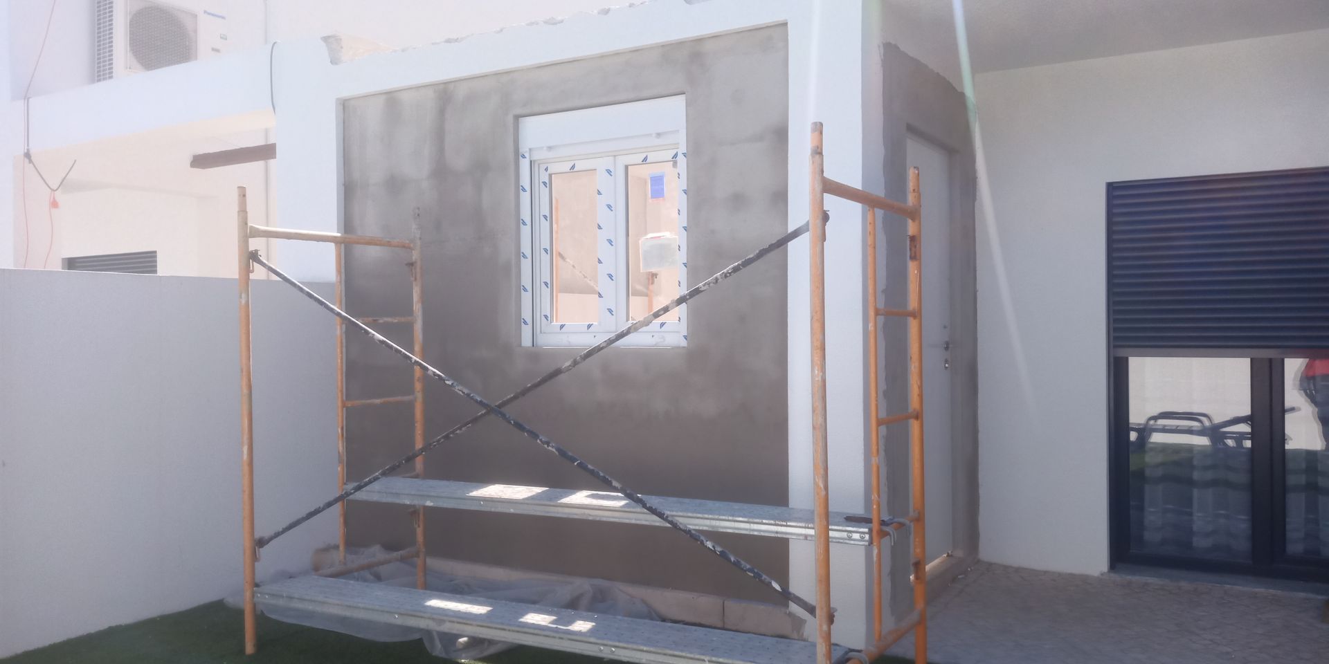 Canteiro de obras com andaimes. Uma janela está no meio de uma parede revestida com gesso cinza.