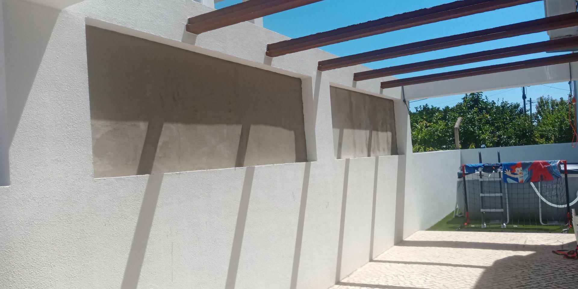 Prédio branco com um pátio sombreado. Ao fundo, uma piscina sob um céu azul.