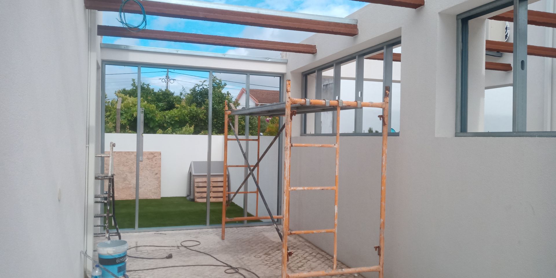 Vista interna de uma obra em construção. Andaimes, janelas e vigas expostas são visíveis. Jardim verde ao fundo.
