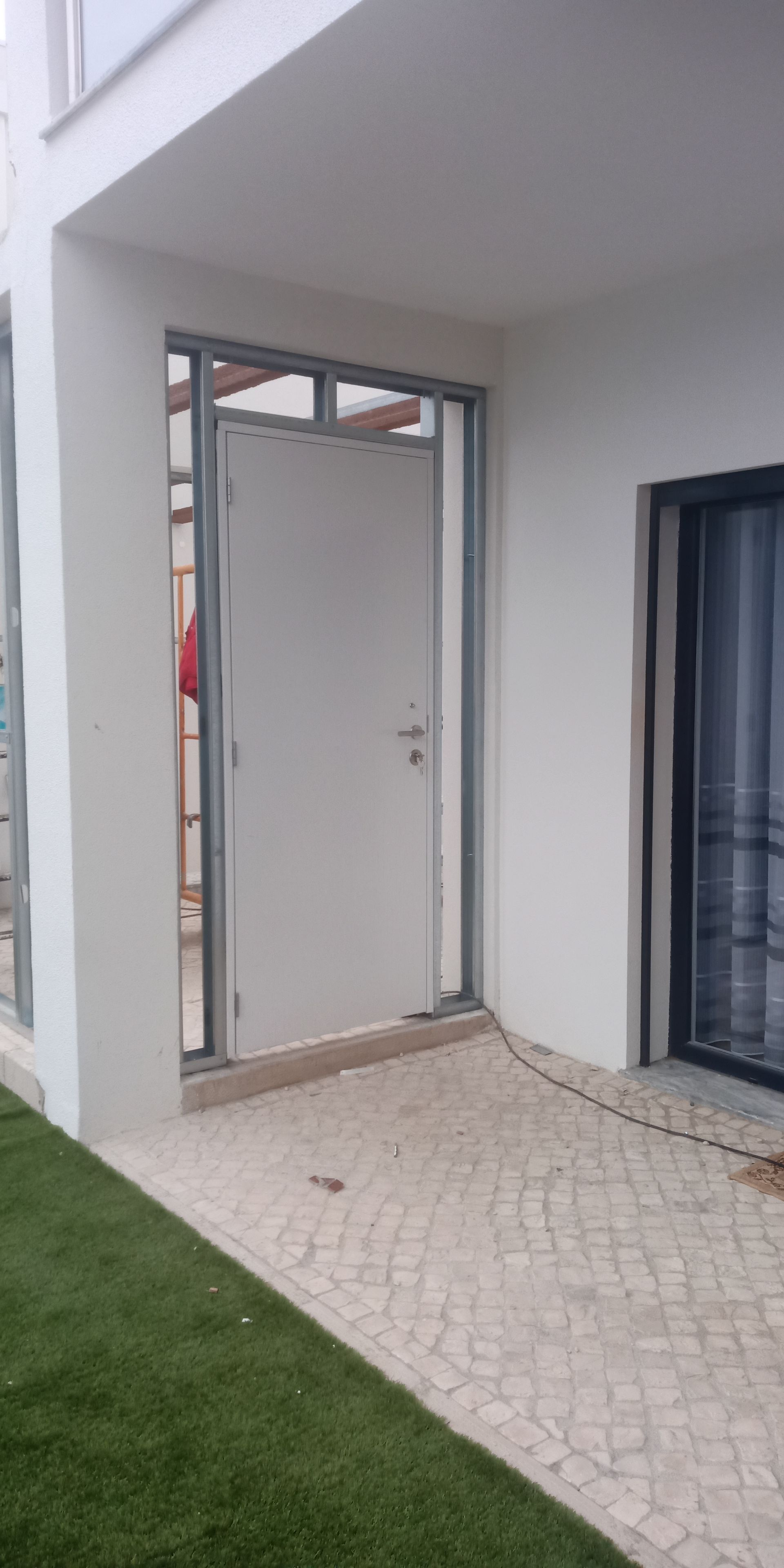 Vista externa de uma porta branca emoldurada em metal. A porta dá para um pátio com piso de pedra e grama artificial.