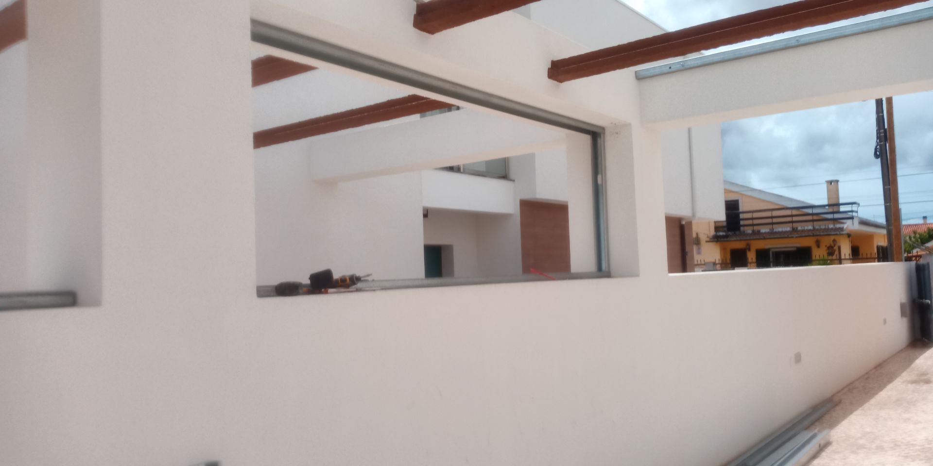 Vista exterior de um edifício branco em construção com vigas de madeira castanhas e vista parcial de uma casa amarela.