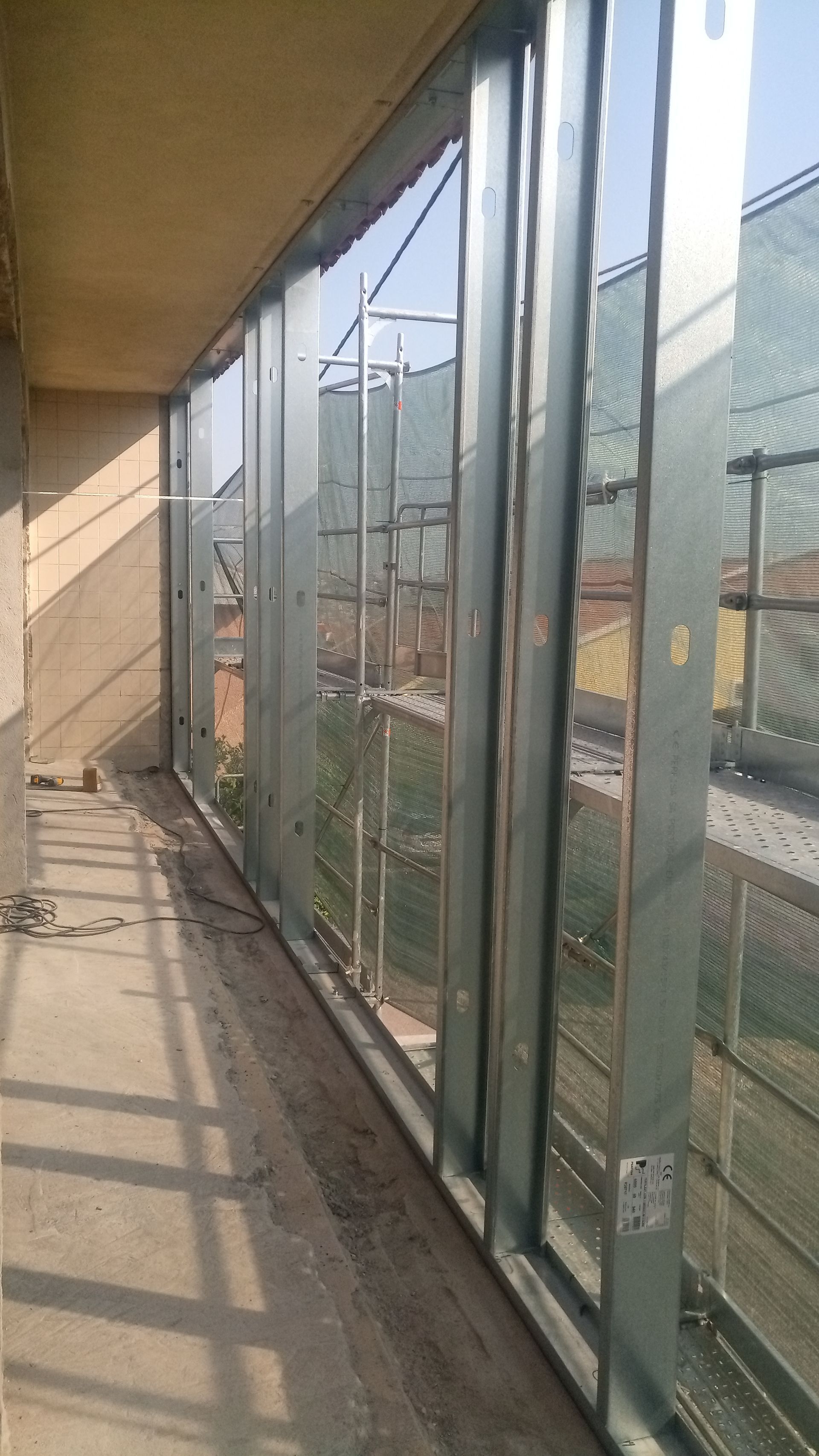 Varanda com painéis de vidro, estrutura metálica e vista para o exterior. Piso de concreto e parede bege.