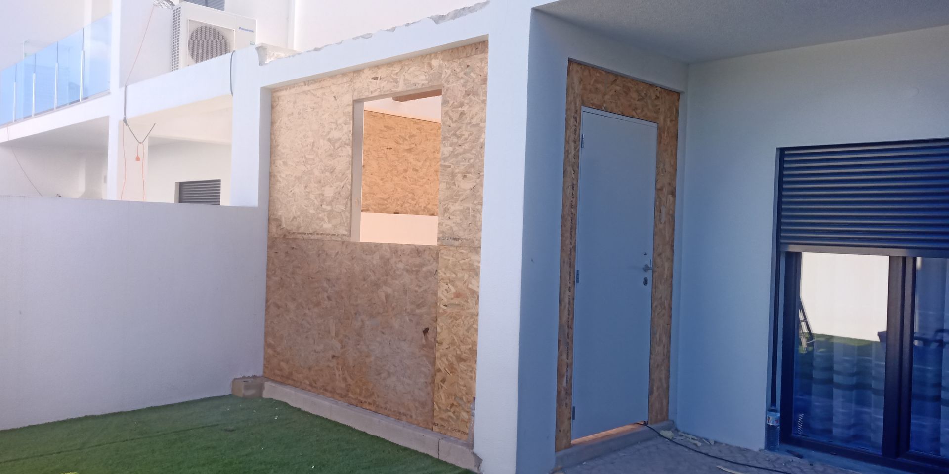 Vista externa de um prédio em construção. As paredes são de cor marrom clara. Há uma porta e uma janela.