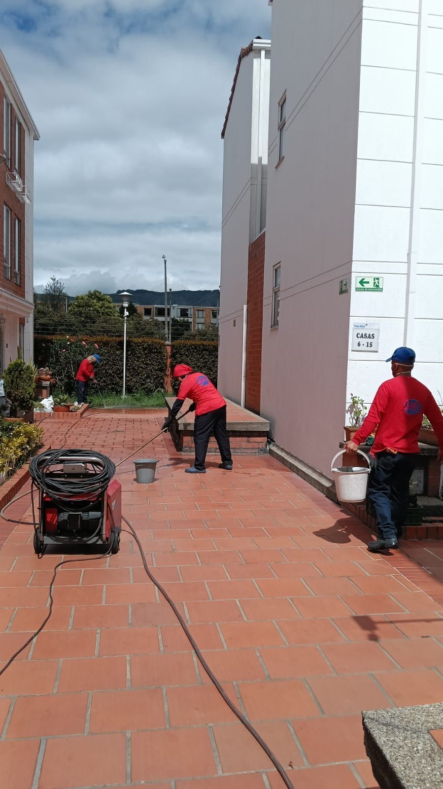Dos trabajadores con chaquetas rojas en una acera de ladrillos, uno empujando un carrito y el otro cargando un cubo junto a un edificio blanco.