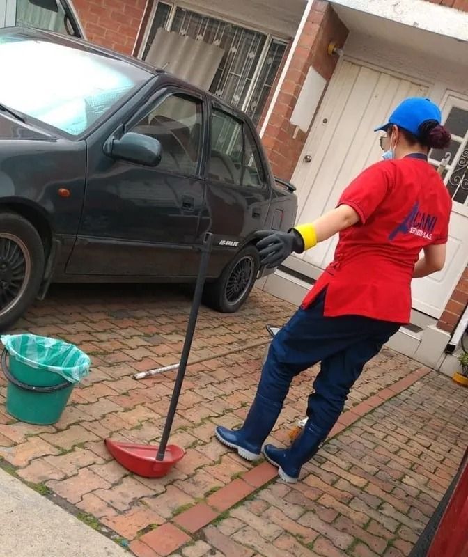Una persona con una camisa roja limpia una entrada de vehículos con una escoba junto a un coche negro aparcado y un cubo verde.