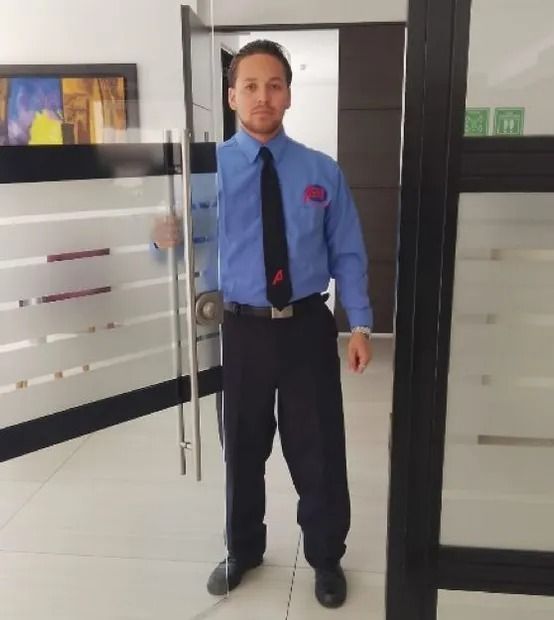 Un hombre con camisa azul y corbata está de pie en el umbral de una puerta, sosteniendo un bastón, en un luminoso pasillo de oficinas.