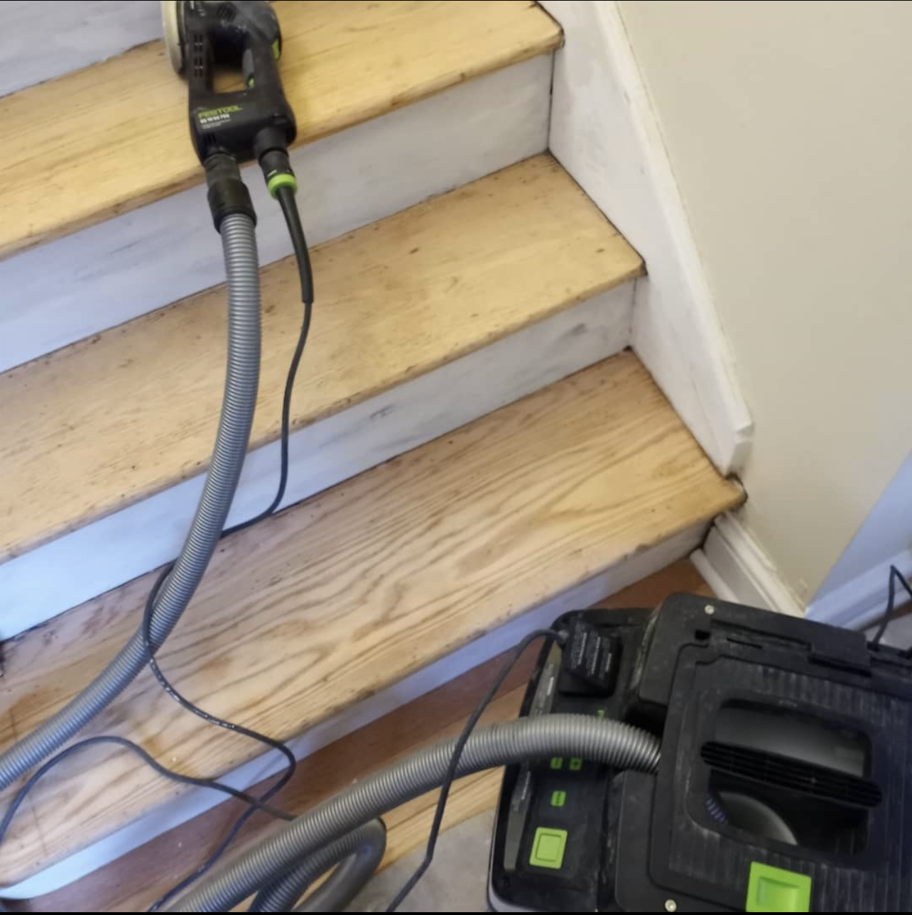 A Festool sander and vacuum connected by a hose sit on a wooden staircase undergoing sanding.