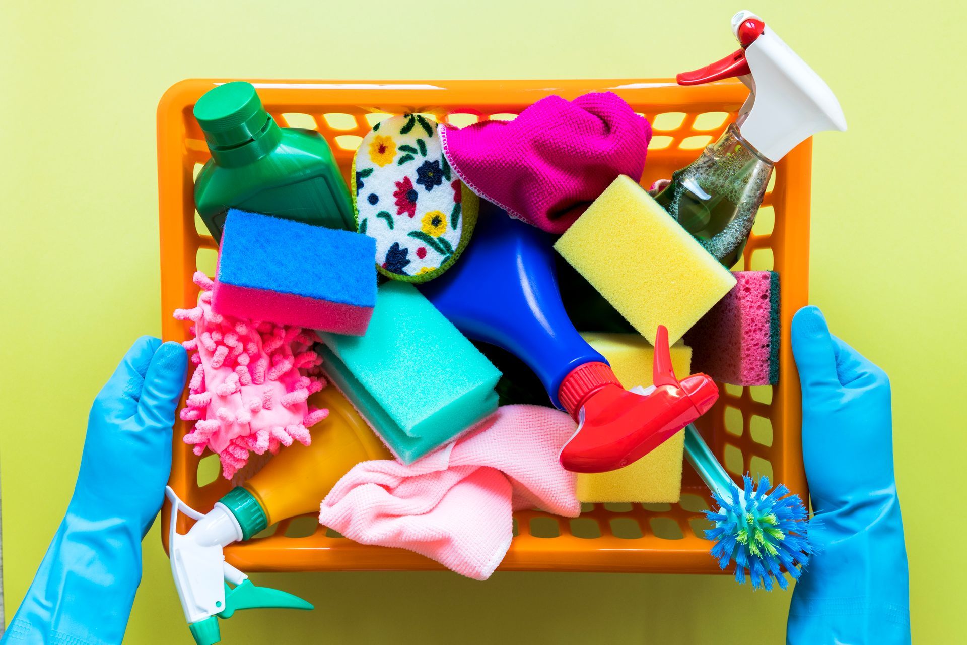 Gloved hands holding a cleaning supply basket with colorful sponges, brushes, and spray bottles on yellow background