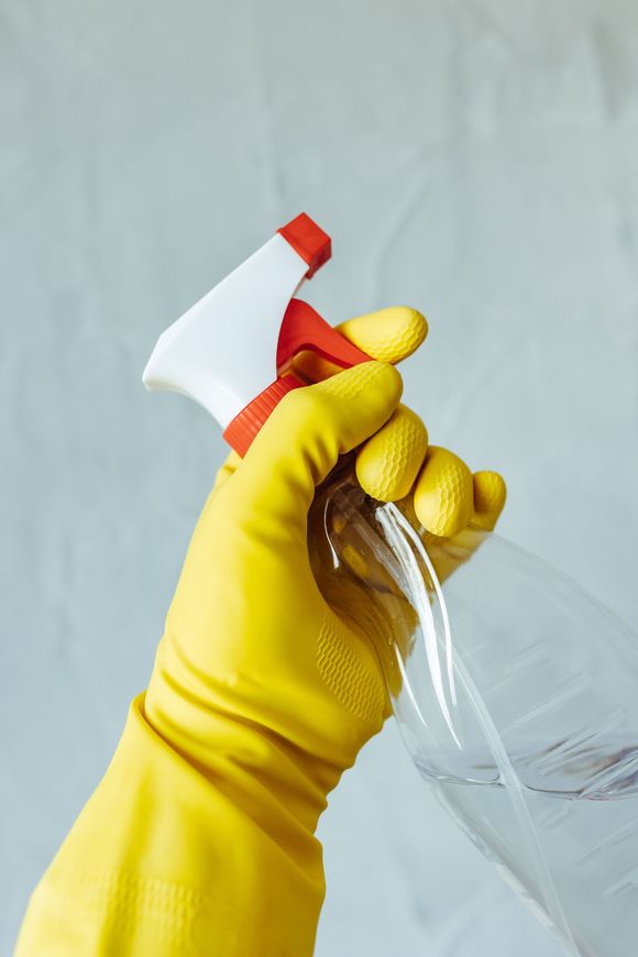 Yellow-gloved hand holding a spray bottle with clear liquid against a pale background