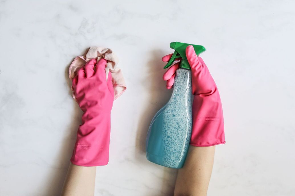 Two gloved hands holding cleaning tools: a cloth and a spray bottle on a light background
