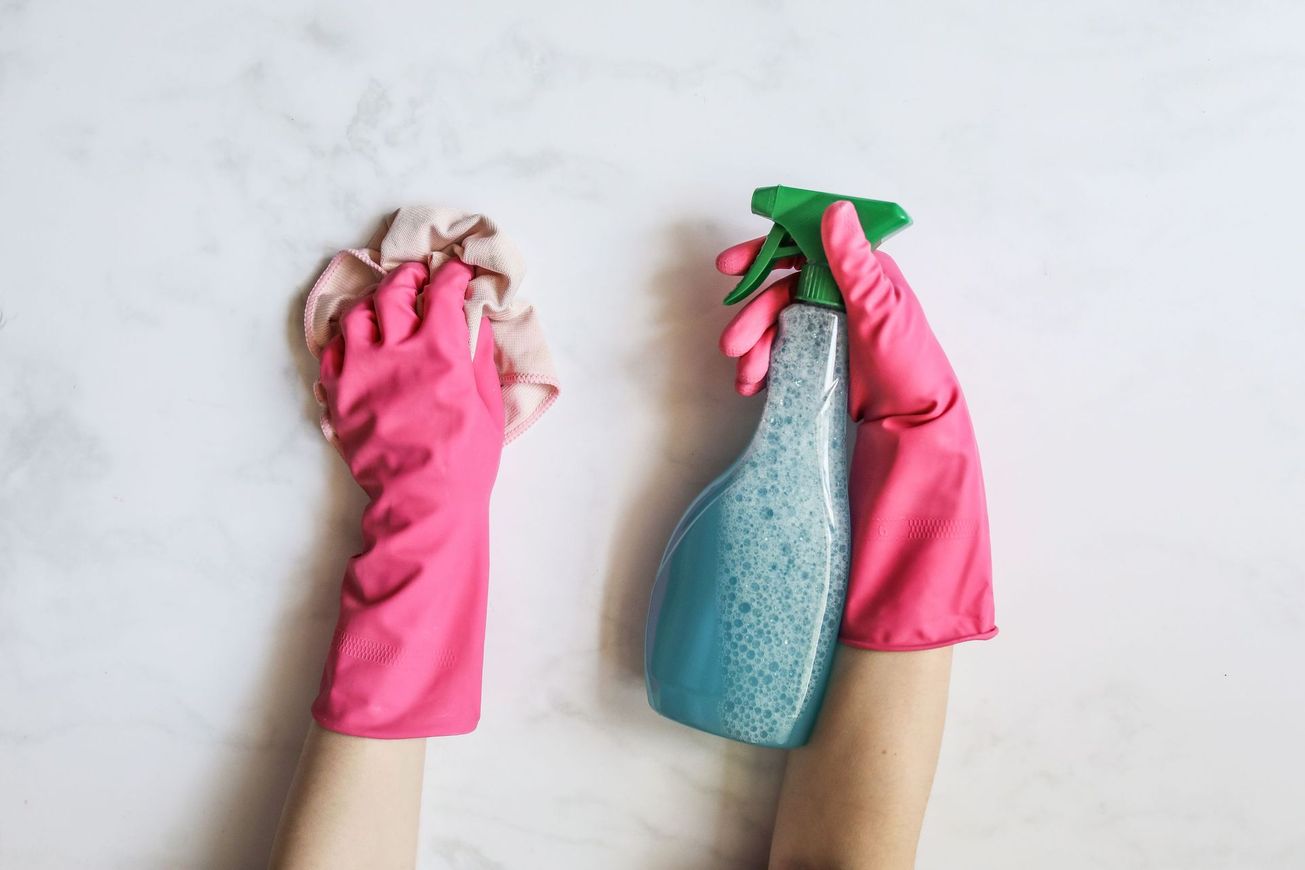 Two gloved hands holding cleaning tools: a cloth and a spray bottle on a light background
