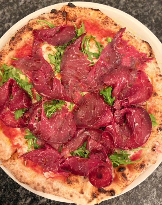 Pizza con salumi e rucola su un piatto bianco. La crosta è dorata, si vedono il sugo rosso e la carne.