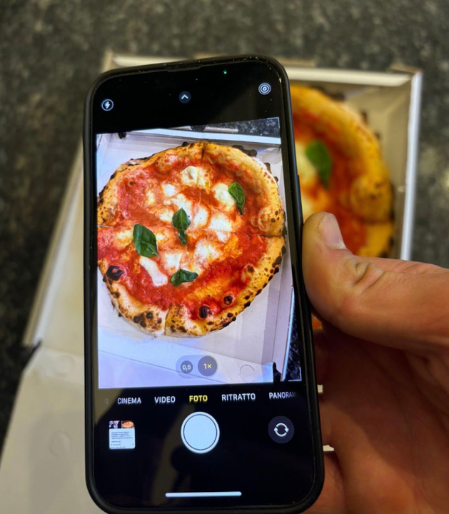 Una mano che tiene un telefono mentre scatta una foto di una pizza Margherita in una scatola. La pizza è condita con salsa rossa, formaggio bianco e basilico.