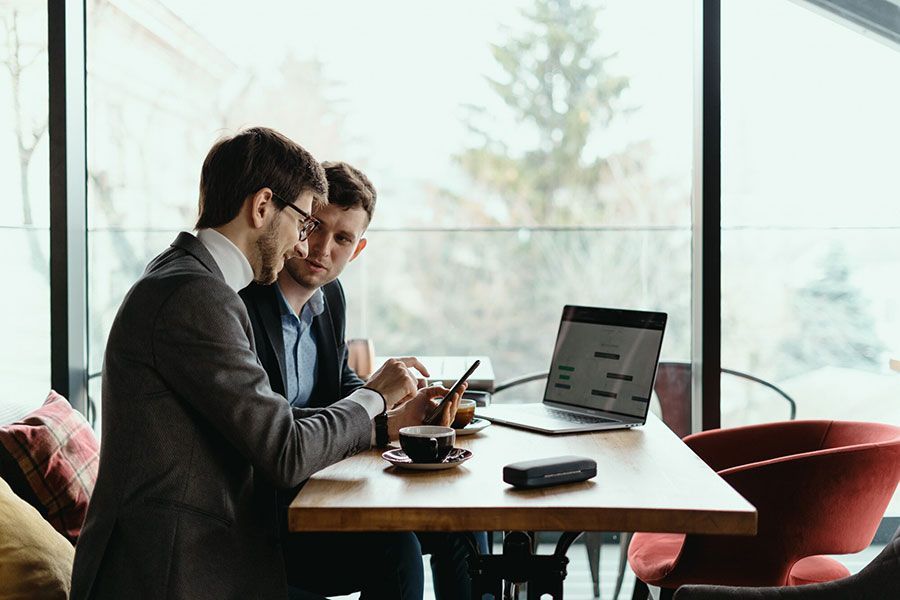 Two colleagues in business attire sit at a wooden table in a sunlit office, looking at a smartphone and an open laptop.