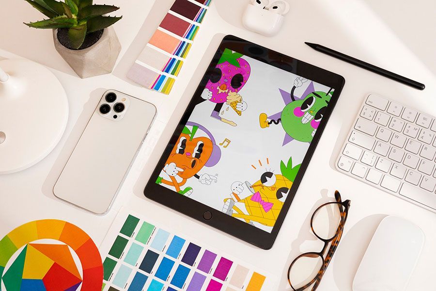 A tablet showing colorful, whimsical fruit characters on a desk with a smartphone, keyboard, glasses, and color swatches.