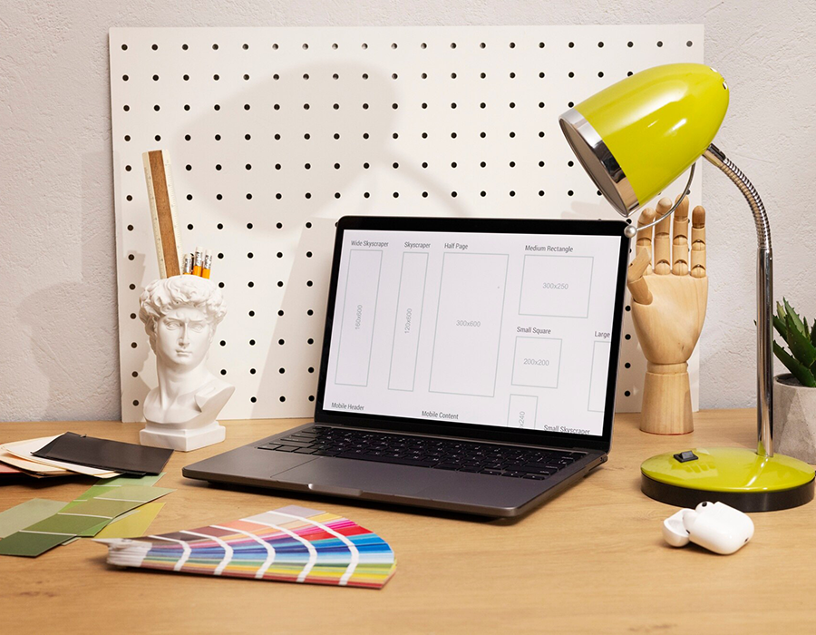 A laptop displaying a wireframe layout on a desk with a bright green lamp, art supplies, and color swatches.