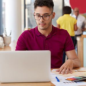 A person in a maroon polo shirt works on a laptop at a desk with papers and a pen, with people blurred in the background.