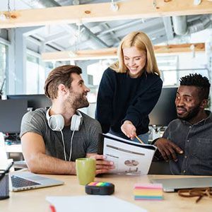 Three colleagues in a modern office space collaborate on a project, reviewing a document together at a wooden table.