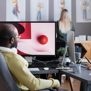 A person in a yellow sweater sits at a computer monitor displaying abstract red shapes, with a colleague working nearby.