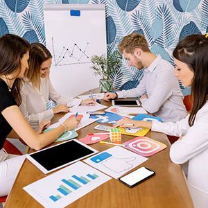 Four people sit at a table collaborating on social media marketing projects featuring app icons and data charts.