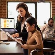 Two professionals in an office collaborate, with one pointing to a document and the other holding a laptop.